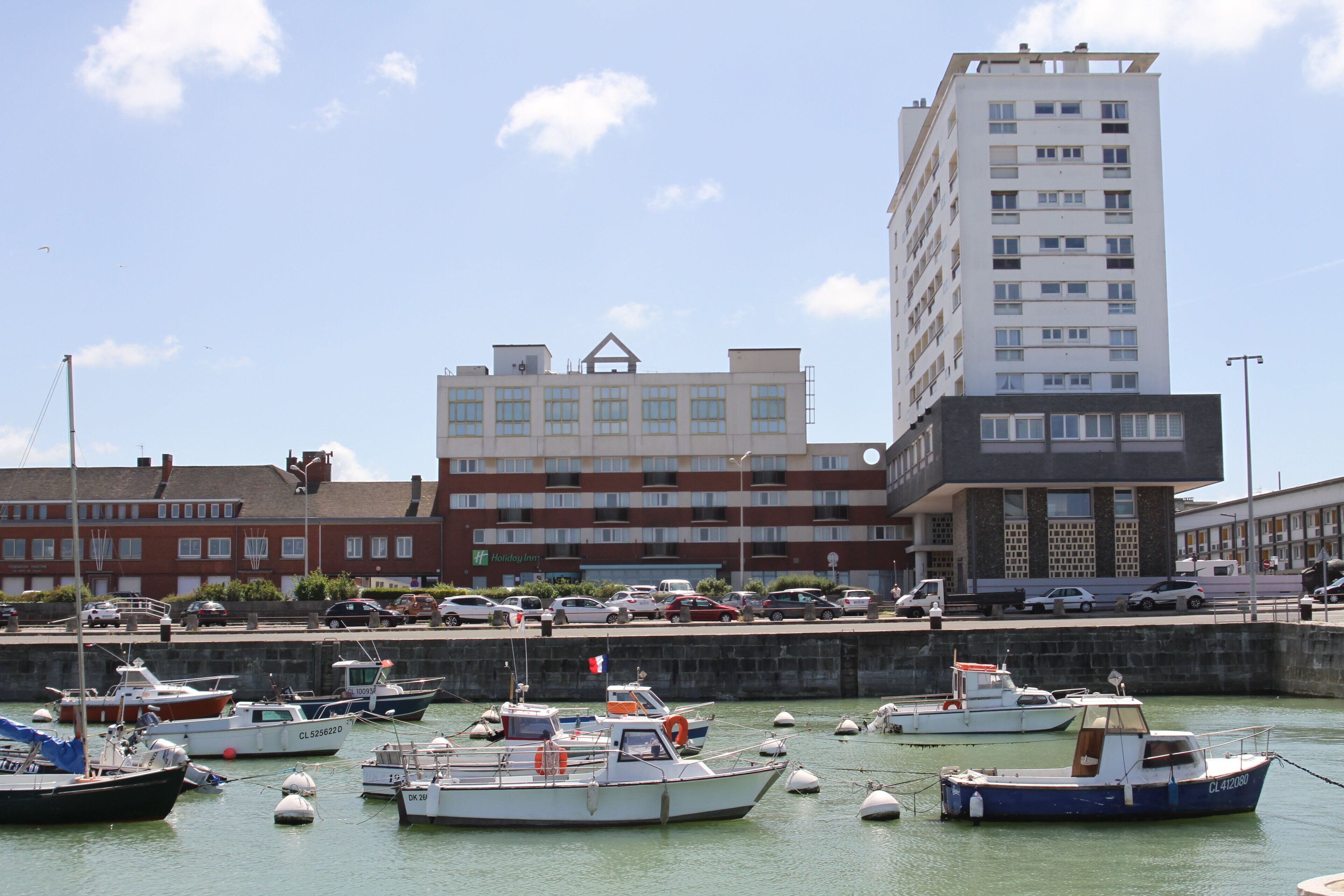 A marina with several boats docked, a tall building with multiple windows, and a parking area with cars.
