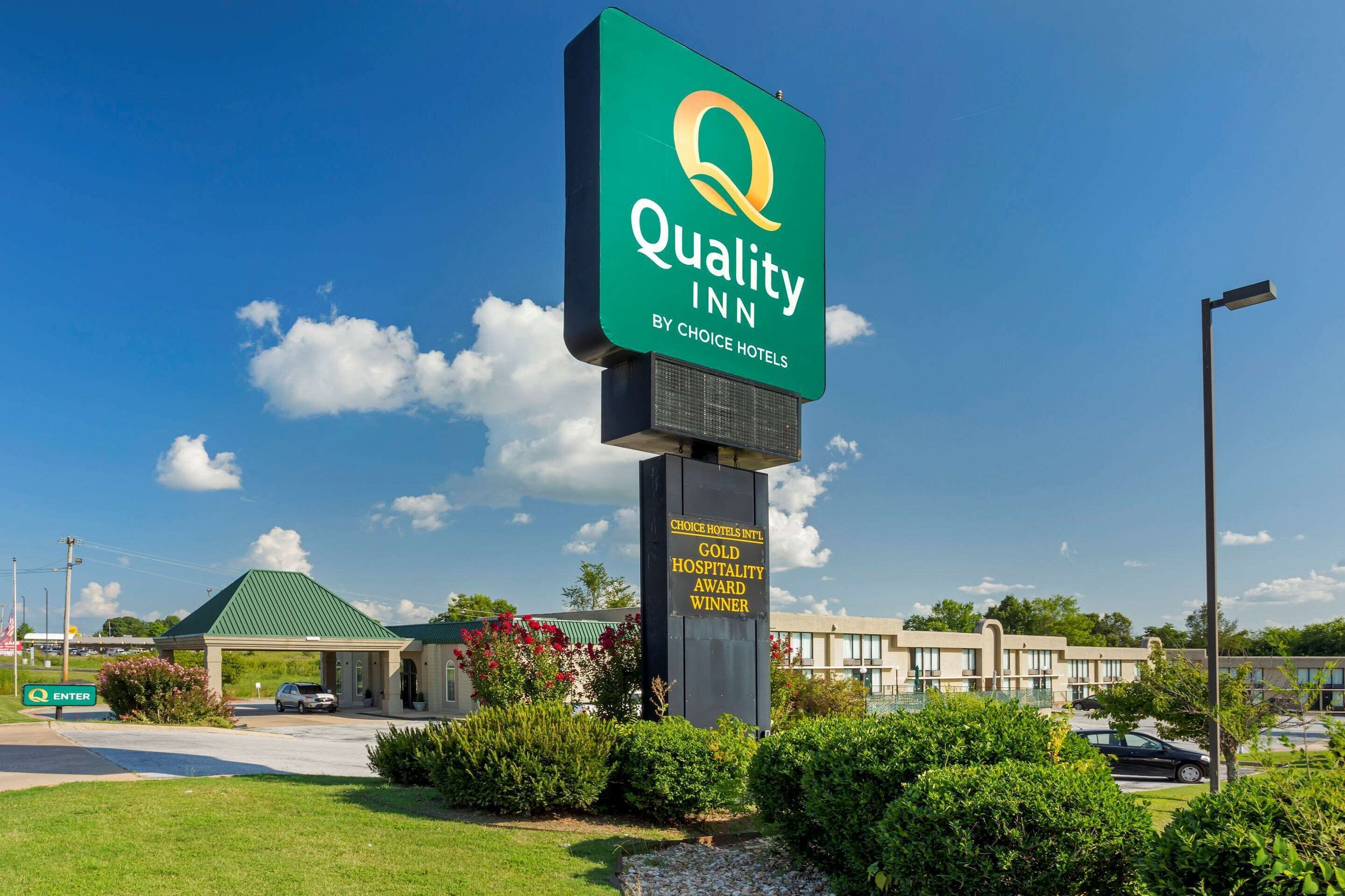 A Quality Inn sign with a green and white logo, a green roofed building, and a clear blue sky.