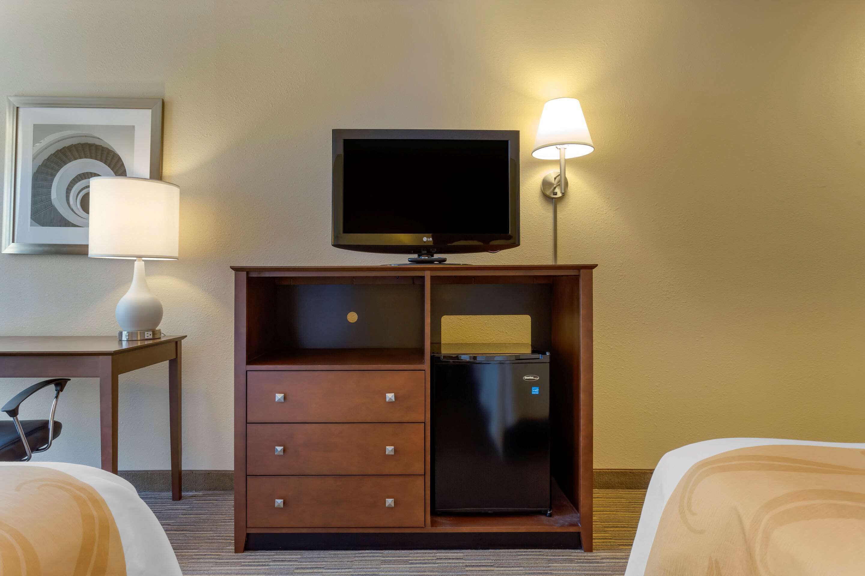 Habitación de hotel con cama, escritorio con lámpara, televisor sobre un soporte y minibar.