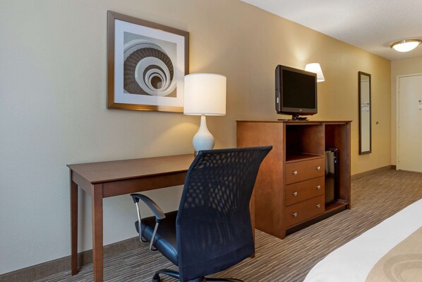 A hotel room with a wooden desk, a black office chair, a white lamp, a television on a wooden cabinet, and a framed artwork on the wall.