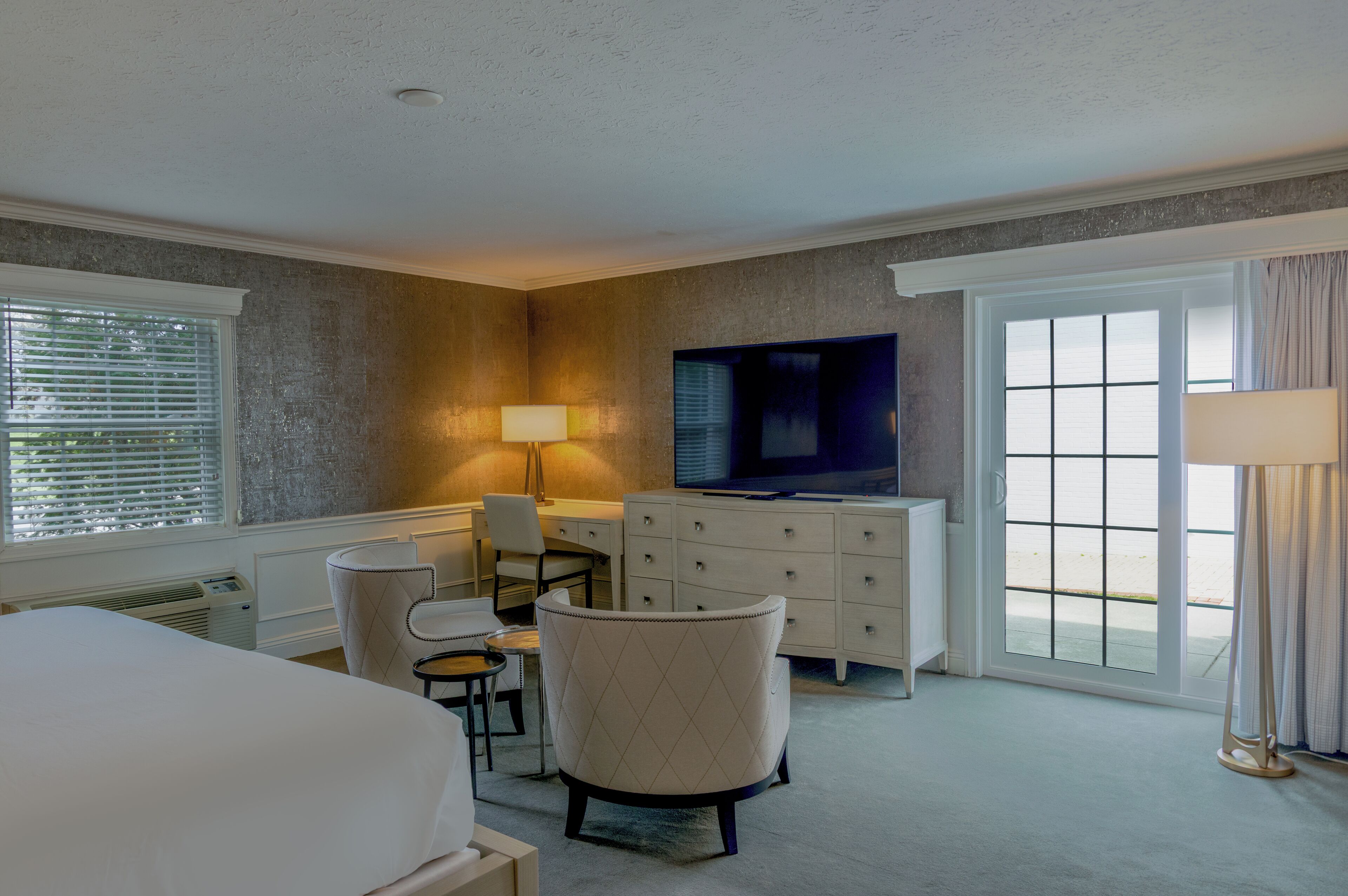 A hotel room with a bed, a desk with a chair, a TV, a dresser, and large windows with curtains.
