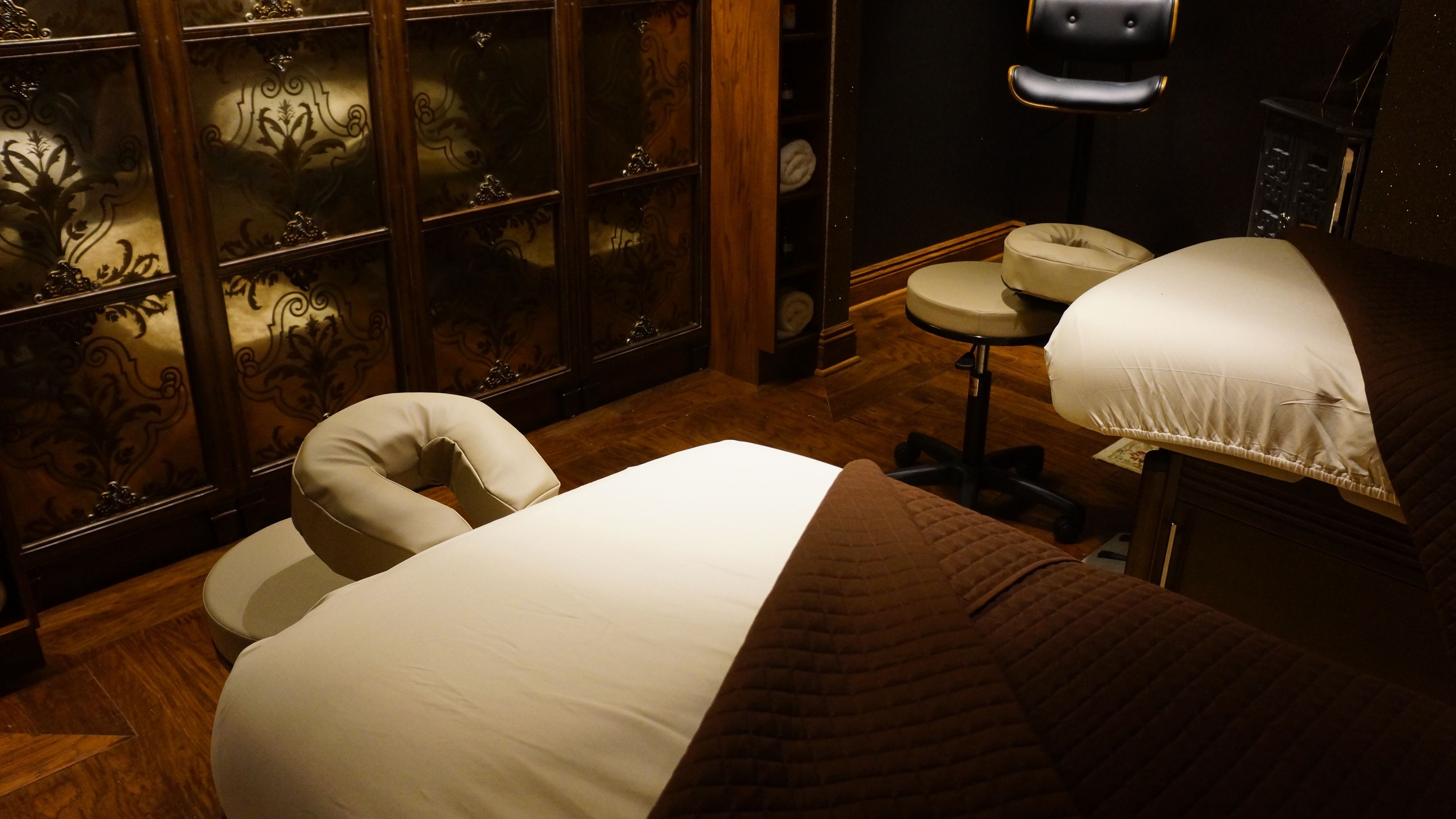 A room with a bed, a massage chair, a wooden cabinet with glass doors, and a small table with a lamp.