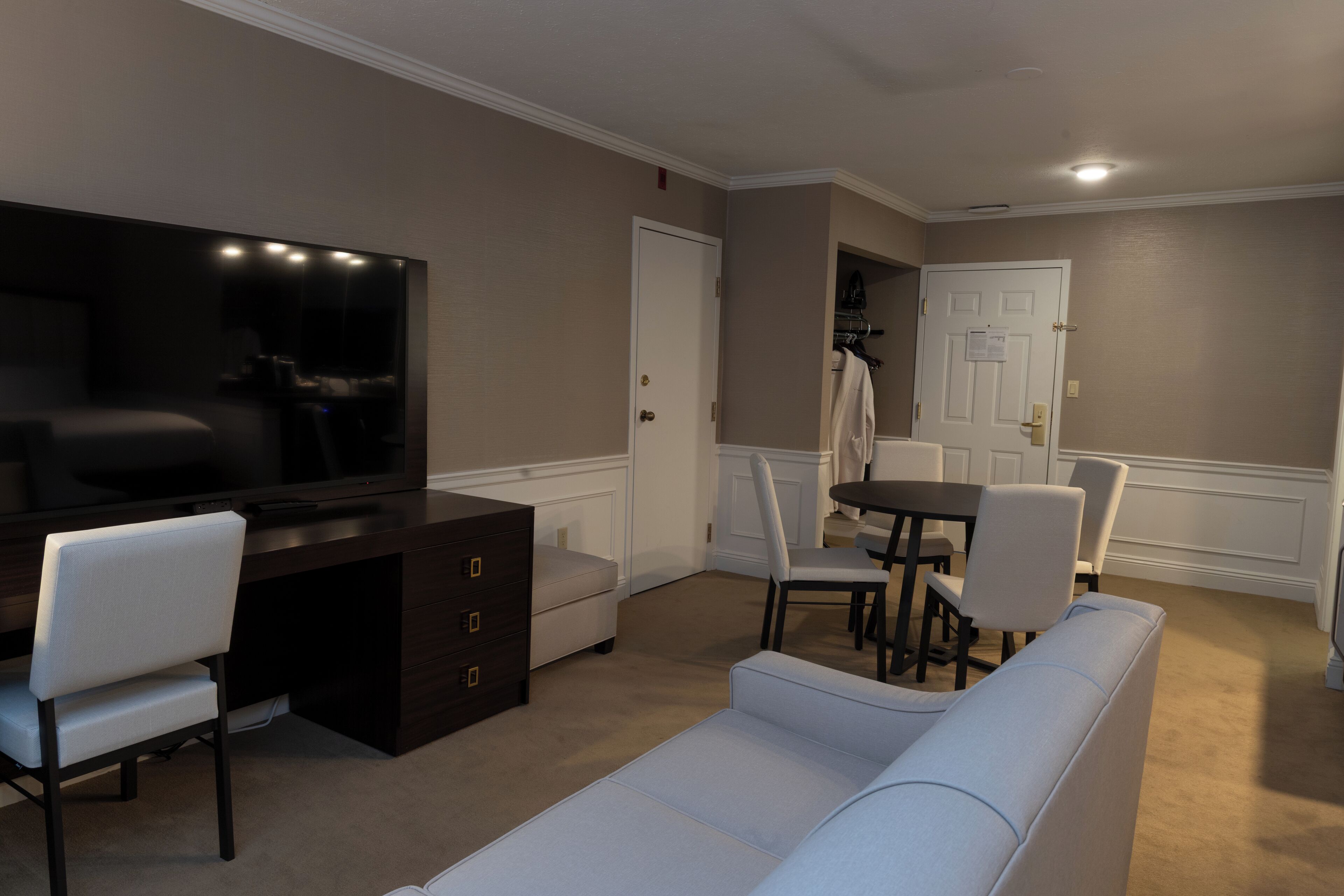 A modern living room with a flat-screen TV, a dark wooden desk, a white sofa, and a dining area with a table and chairs.