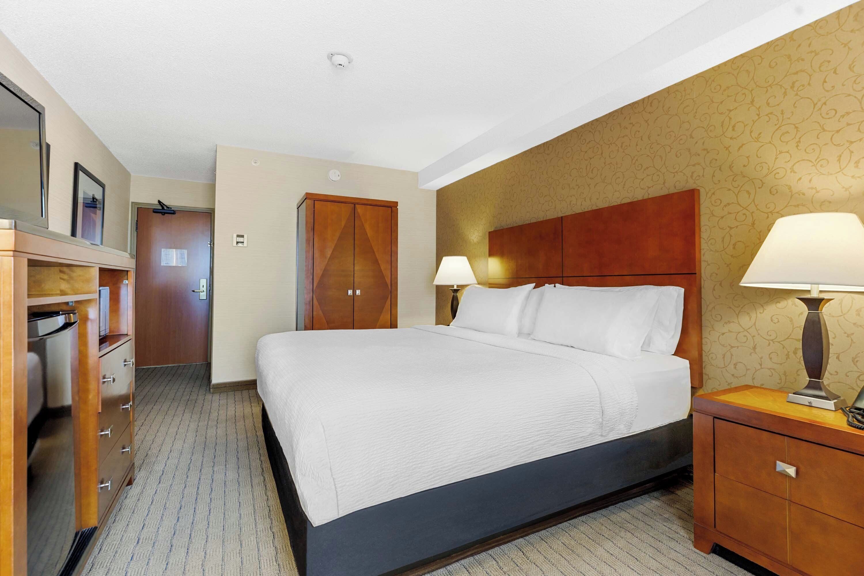 Ein Hotelzimmer mit einem großen Bett, einem Holzkopfstück, zwei Nachttischlampen, einem Kleiderschrank und einer Tür zu einem weiteren Raum.