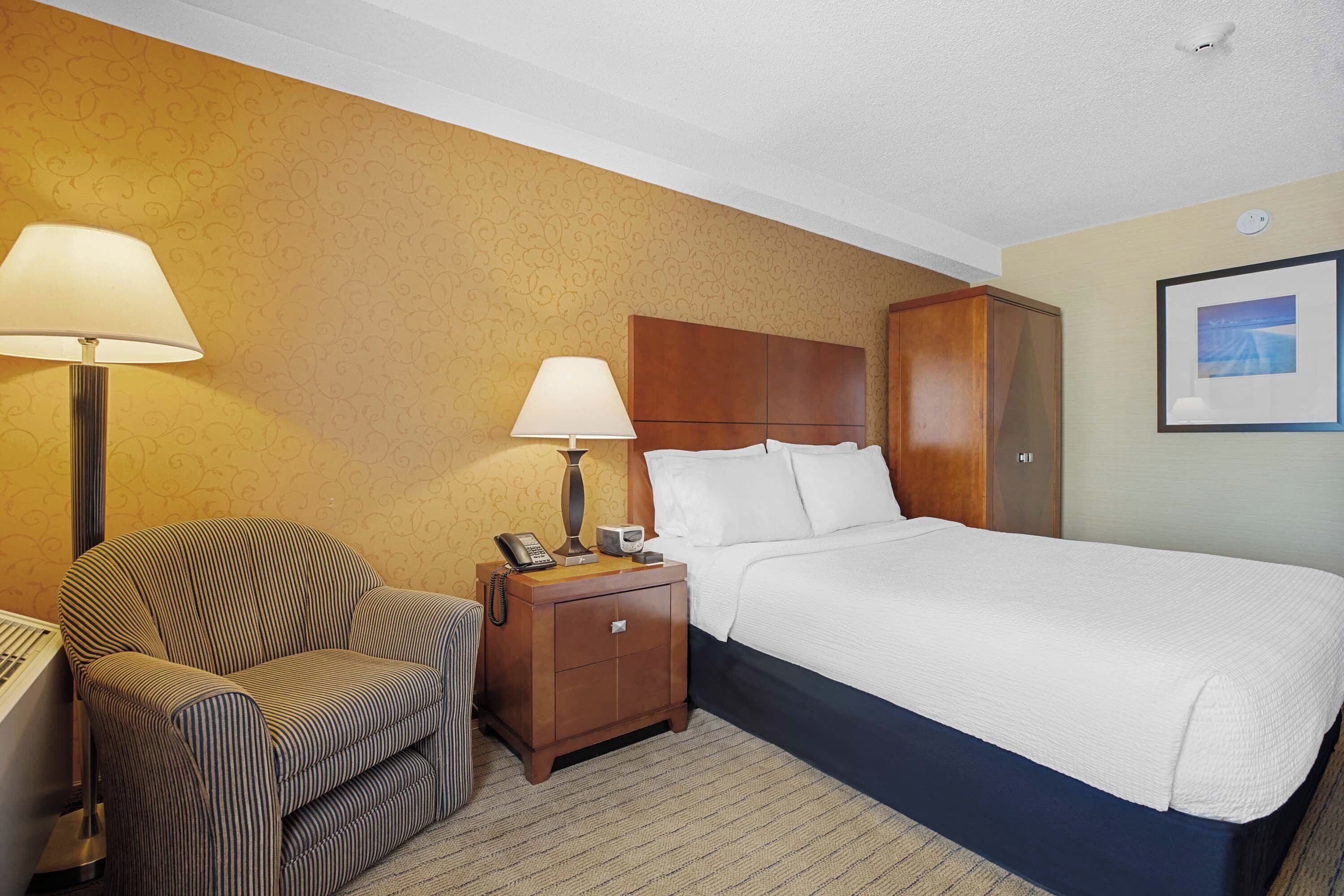 A hotel room with a bed, a nightstand with a lamp, a chair, and a wardrobe.