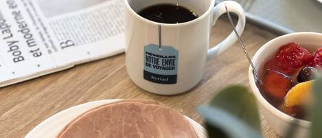 Ein Frühstückstisch mit Zeitung, Tasse, Glas Orangensaft, Teller mit Schinken, Käse und Pfannkuchen sowie einer Obstschale.