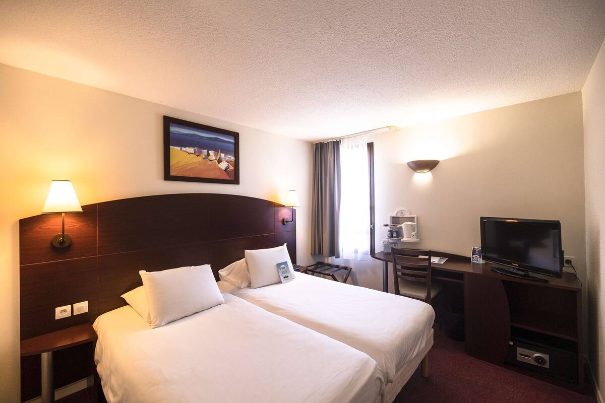 A hotel room with two beds, a desk with a television, a coffee machine, and a painting on the wall.