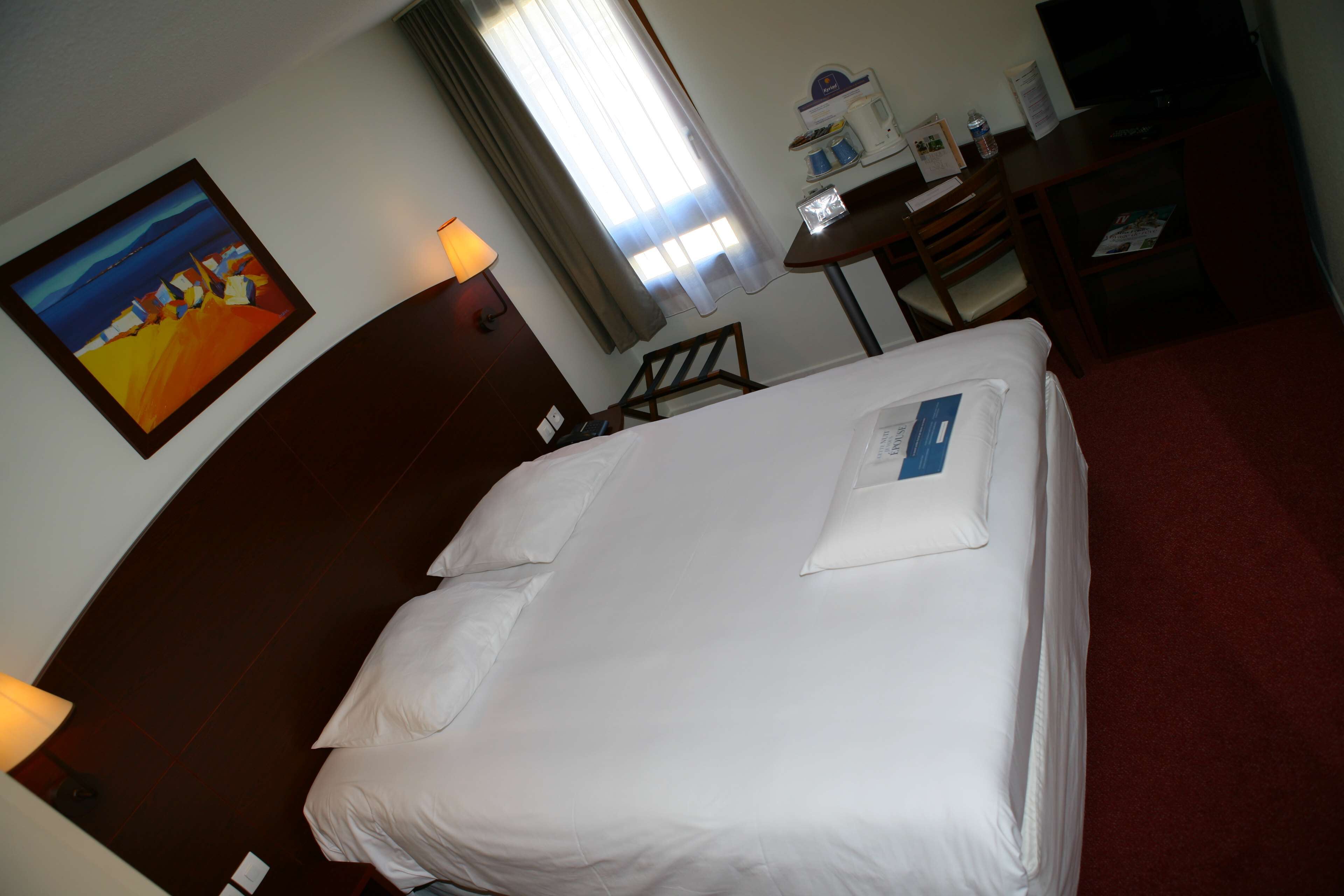 A hotel room with a neatly made bed, a desk with a chair, a television, and a painting on the wall.