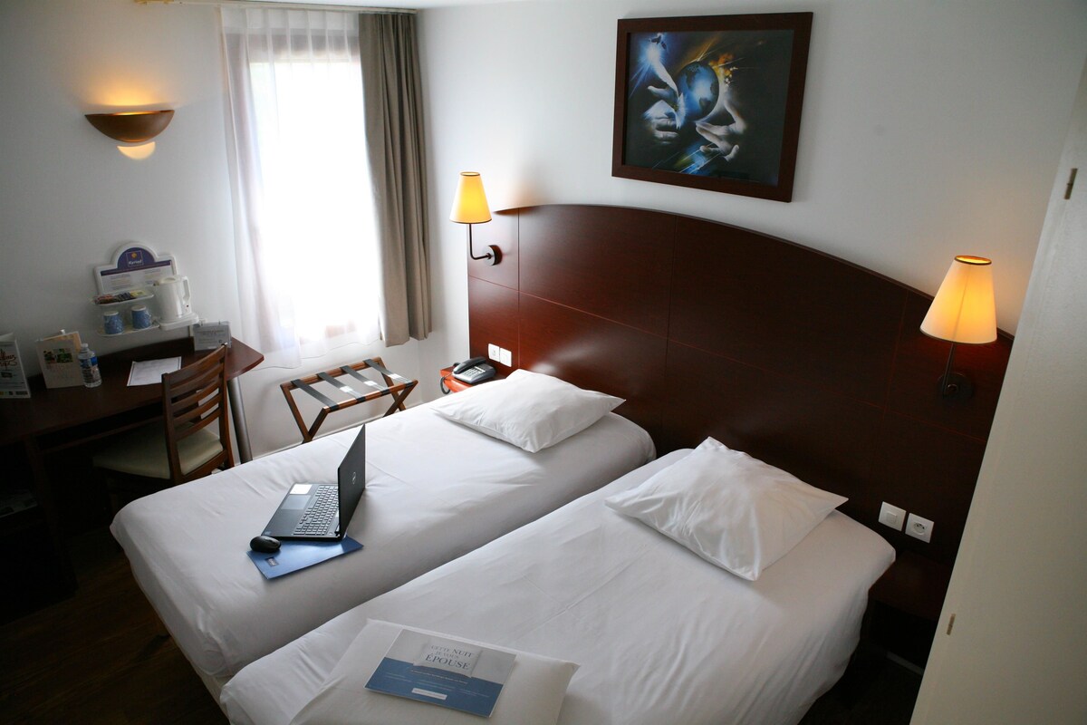 A hotel room with two beds, a desk with a laptop, a chair, and a painting on the wall.