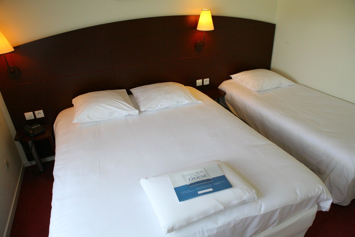 A hotel room with two beds, each with white linens and pillows. There is a nightstand with a lamp and a phone. A folded towel with a hotel name and logo is on the bed.