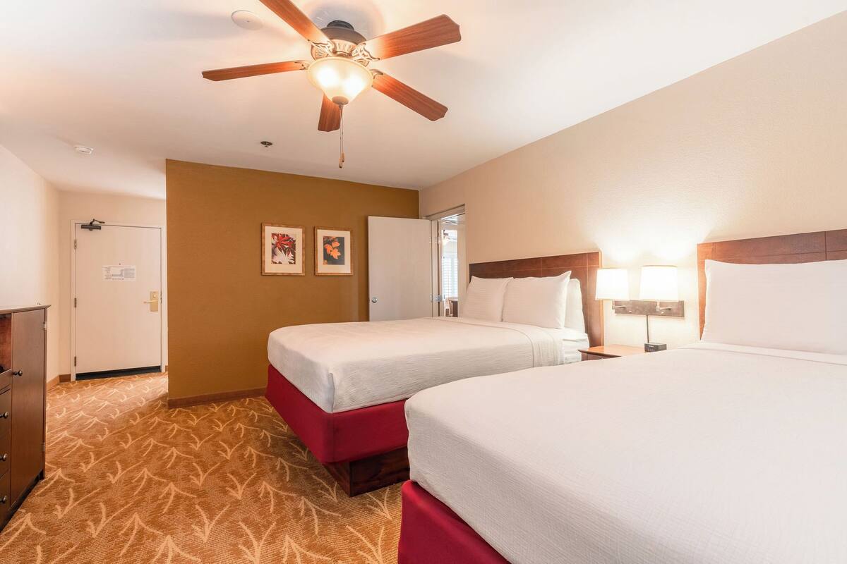 A hotel room with two beds, a ceiling fan, a door, and a carpeted floor.