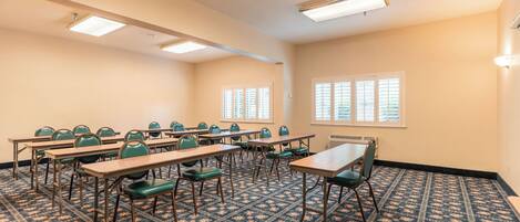 A conference room with multiple tables and chairs, large windows with blinds, and patterned carpet.