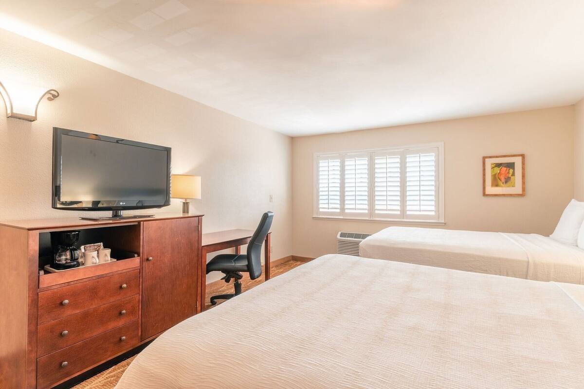 A hotel room with a TV, desk, chair, two beds, and a window with blinds.
