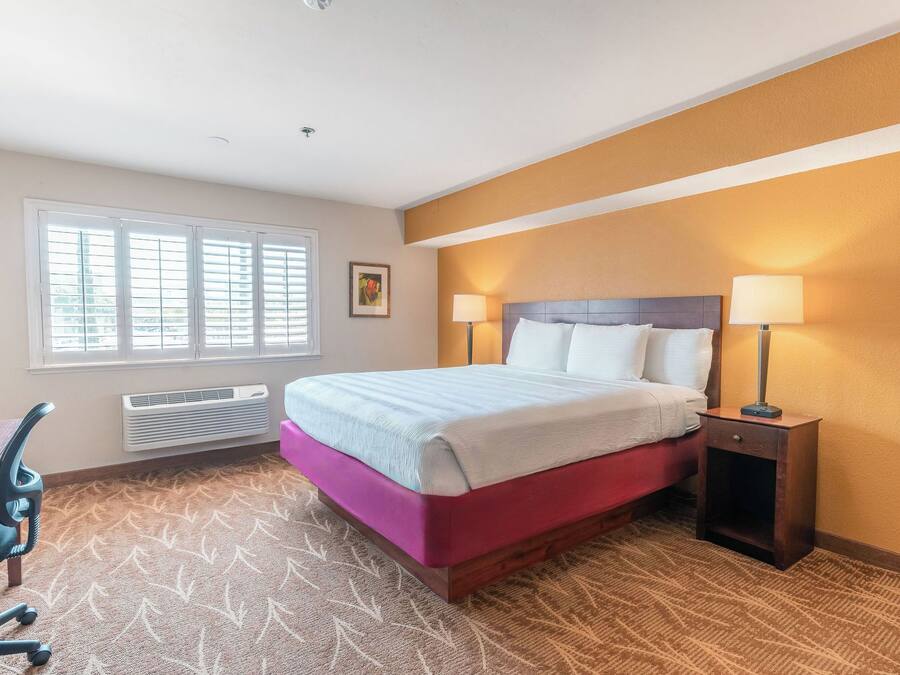 Une chambre d’hôtel comprenant un lit, deux lampes de chevet, un bureau avec une chaise et une fenêtre avec des stores.