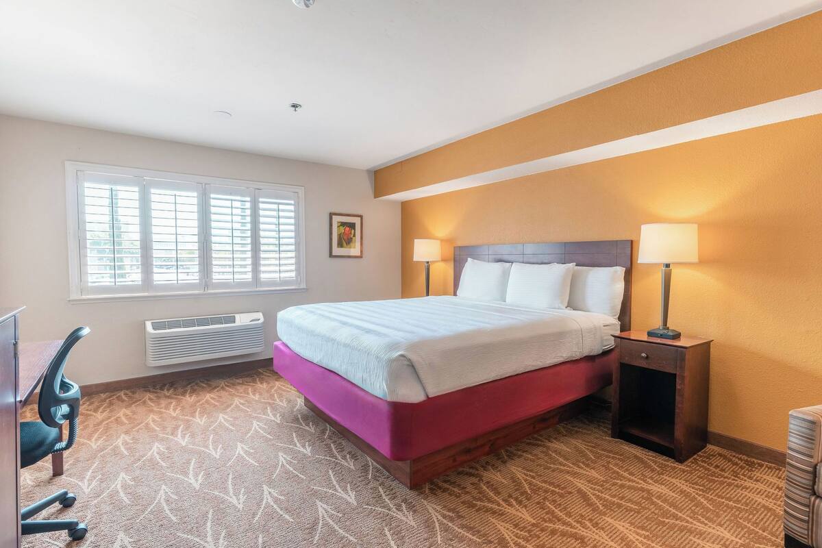 A hotel room with a bed, two bedside lamps, a desk with a chair, and a window with blinds.