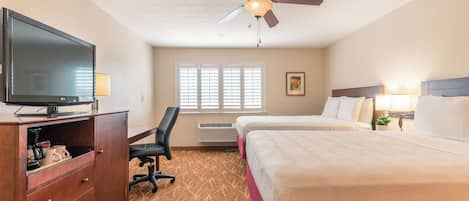 Quarto de hotel com televisão de tela plana, uma escrivaninha com cadeira, duas camas, ventilador de teto e janela com persianas.