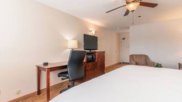 A hotel room with a bed, a desk with a chair, a television, a microwave, a ceiling fan, and a door.