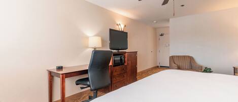 A hotel room with a bed, a desk with a chair, a television, a microwave, a ceiling fan, and a door.