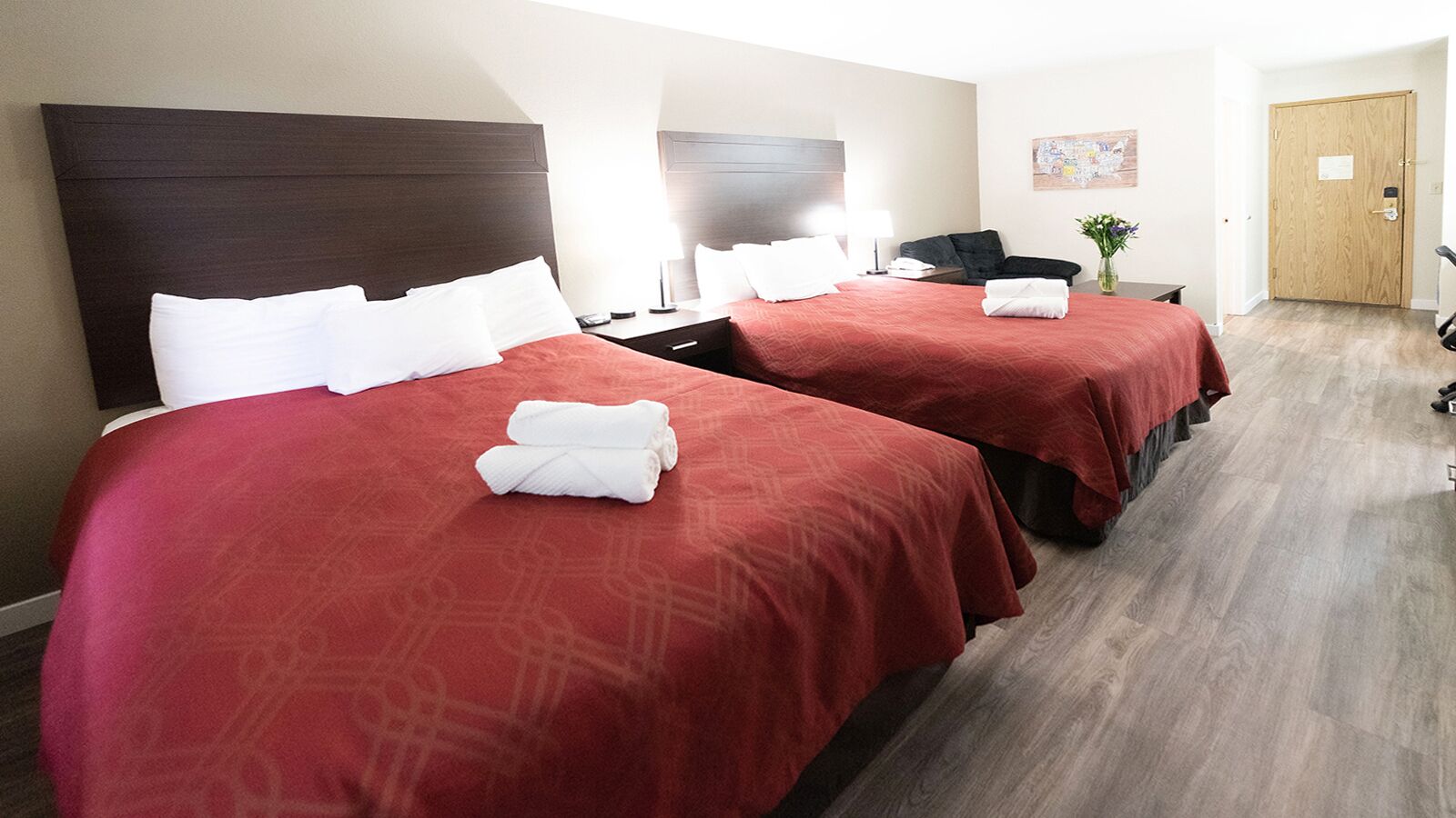 A hotel room with two beds, each with a red bedspread and white pillows, a wooden headboard, a bedside table with a lamp, and a wooden door.