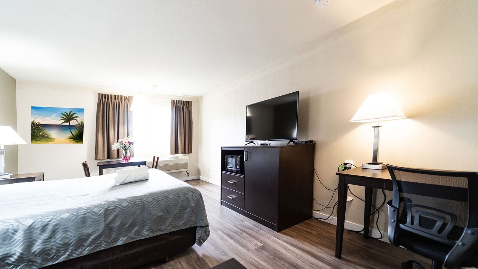 Et hotelværelse med en seng, et skrivebord med lampe, et fjernsyn og et maleri på væggen.