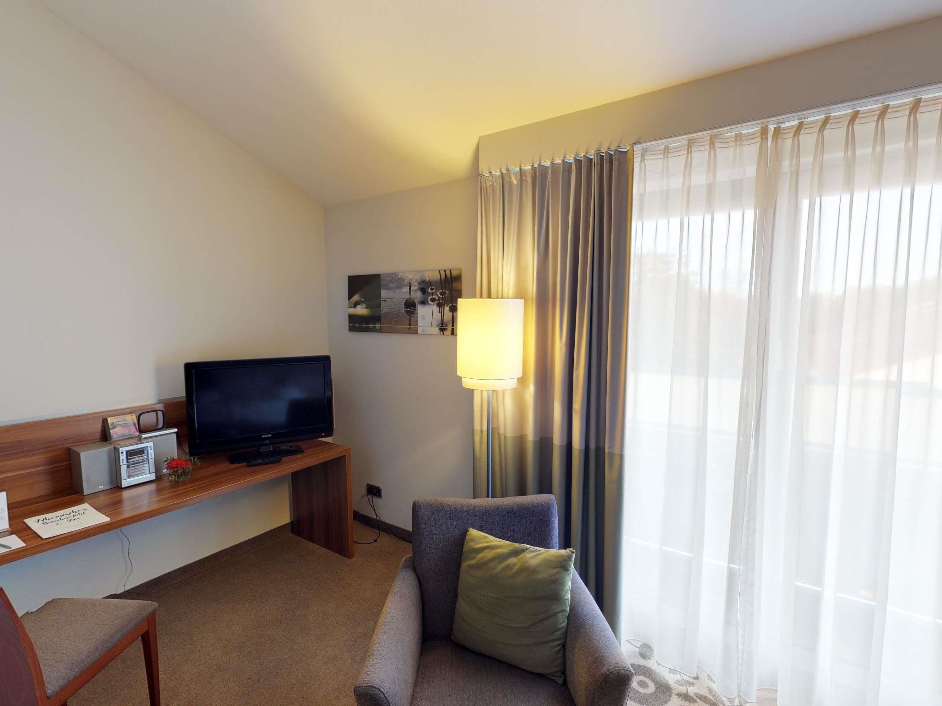 Habitació d'hotel amb televisió de pantalla plana, taula petita amb ràdio i un llibre, sofà amb almoçó i finestra gran amb cortines translúcides.