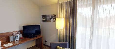 Habitació d'hotel amb televisió de pantalla plana, taula petita amb ràdio i un llibre, sofà amb almoçó i finestra gran amb cortines translúcides.
