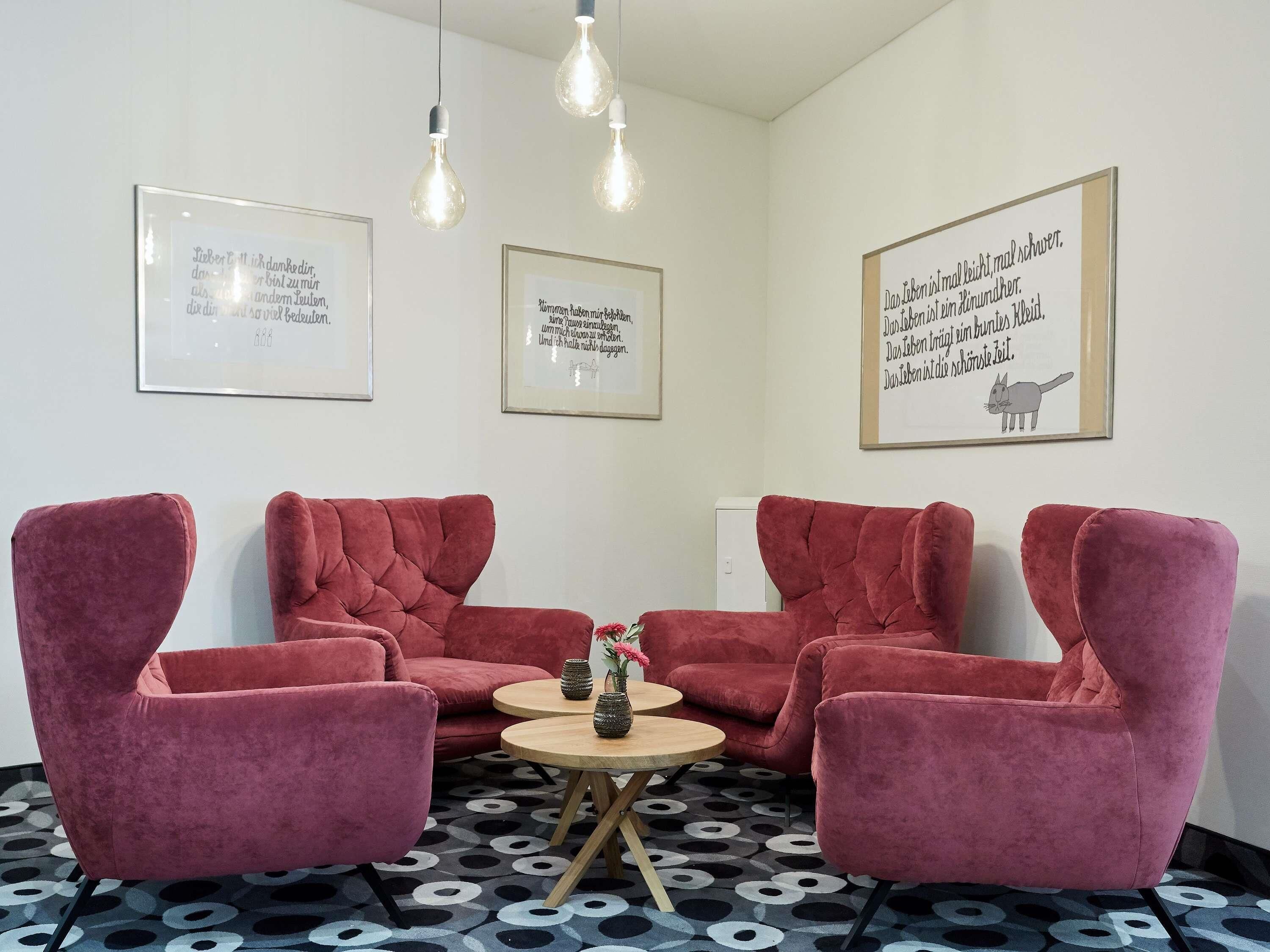 A room with two red velvet armchairs, a small wooden table with vases, and three wall-mounted light fixtures.