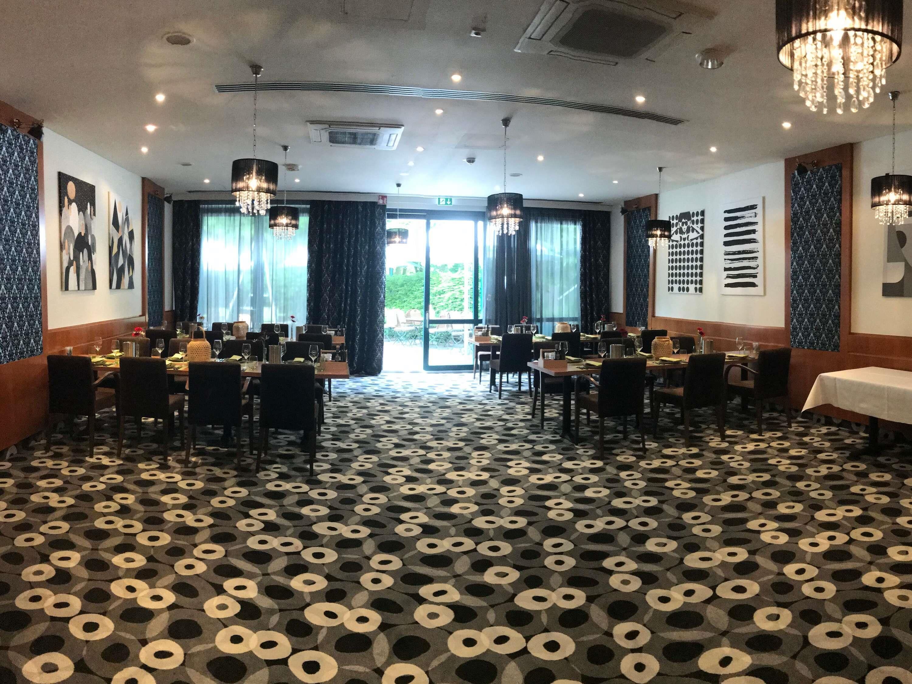 A hotel dining area with tables set for guests, large windows with curtains, and a patterned floor.