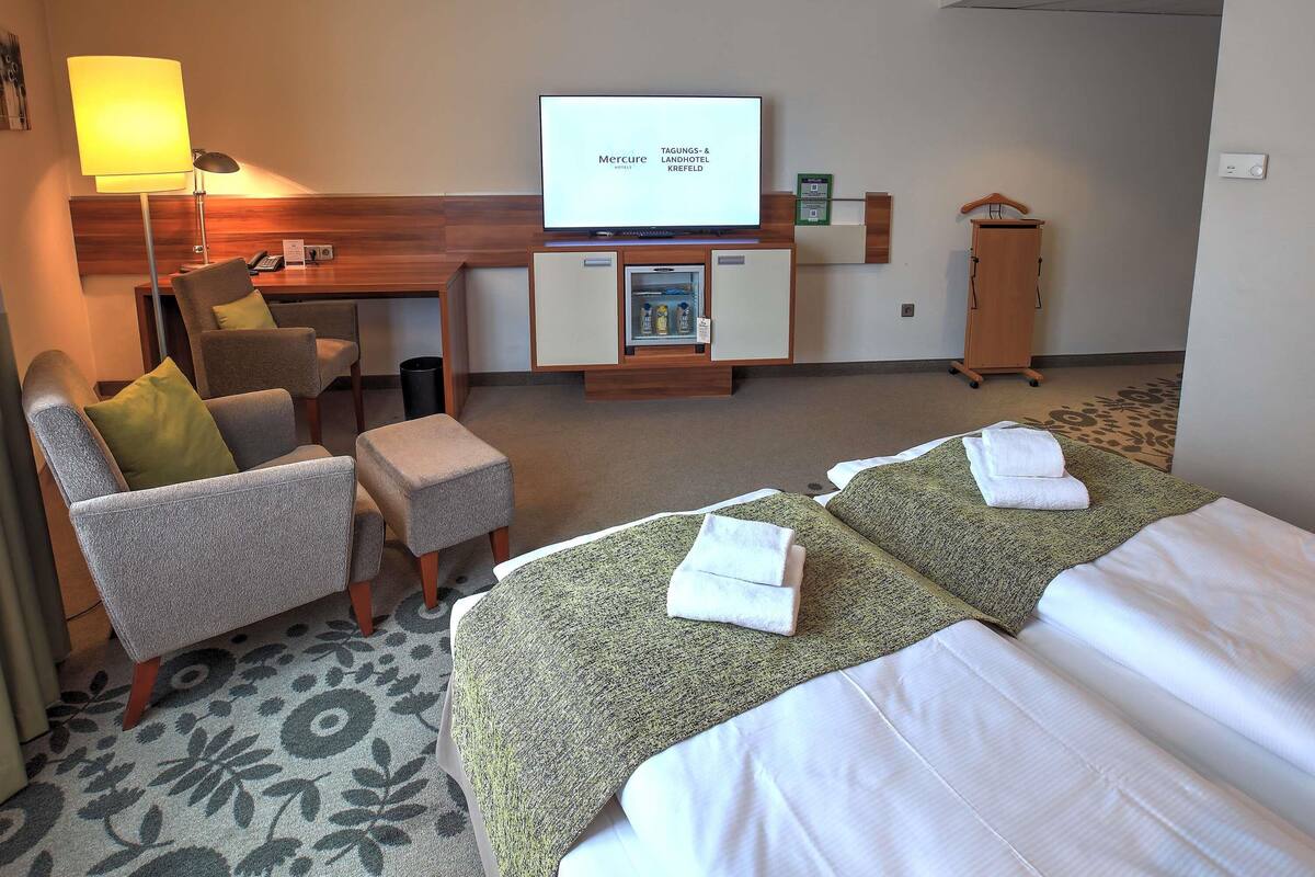 Habitación de hotel con una cama, dos sillones, un escritorio con ordenador, televisión y un mueble pequeño.
