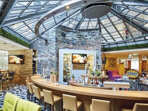 A bar area with a stone wall, a high ceiling with a glass roof, and a seating area with chairs and a sofa. - Mercure Tagungs- & Landhotel Krefeld (Krefeld)