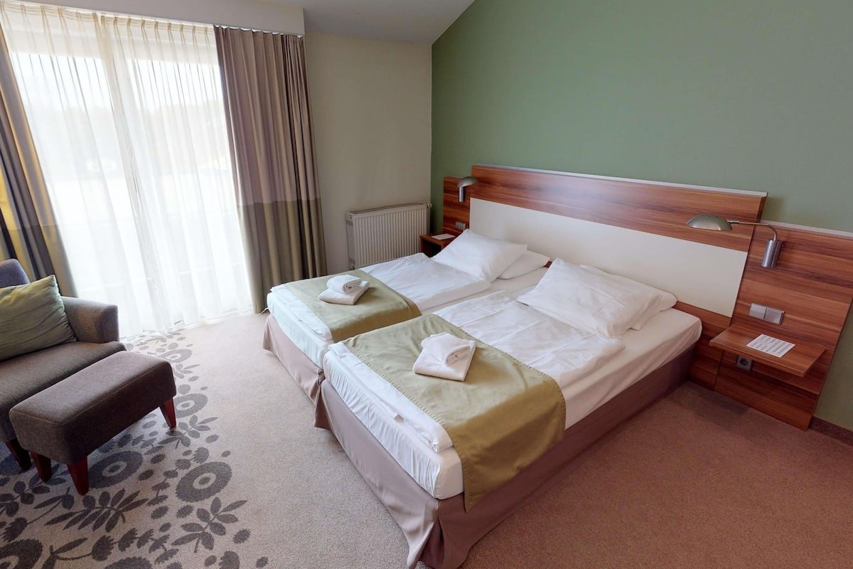 A hotel room with two beds, a chair, a radiator, and a window with curtains.