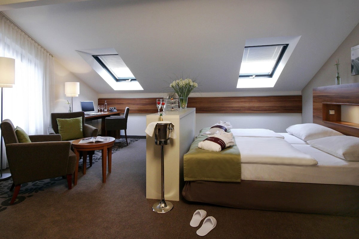 Habitación de hotel con cama, escritorio y dos sillas. Hay tragaluces y una pequeña mesa con un florero.