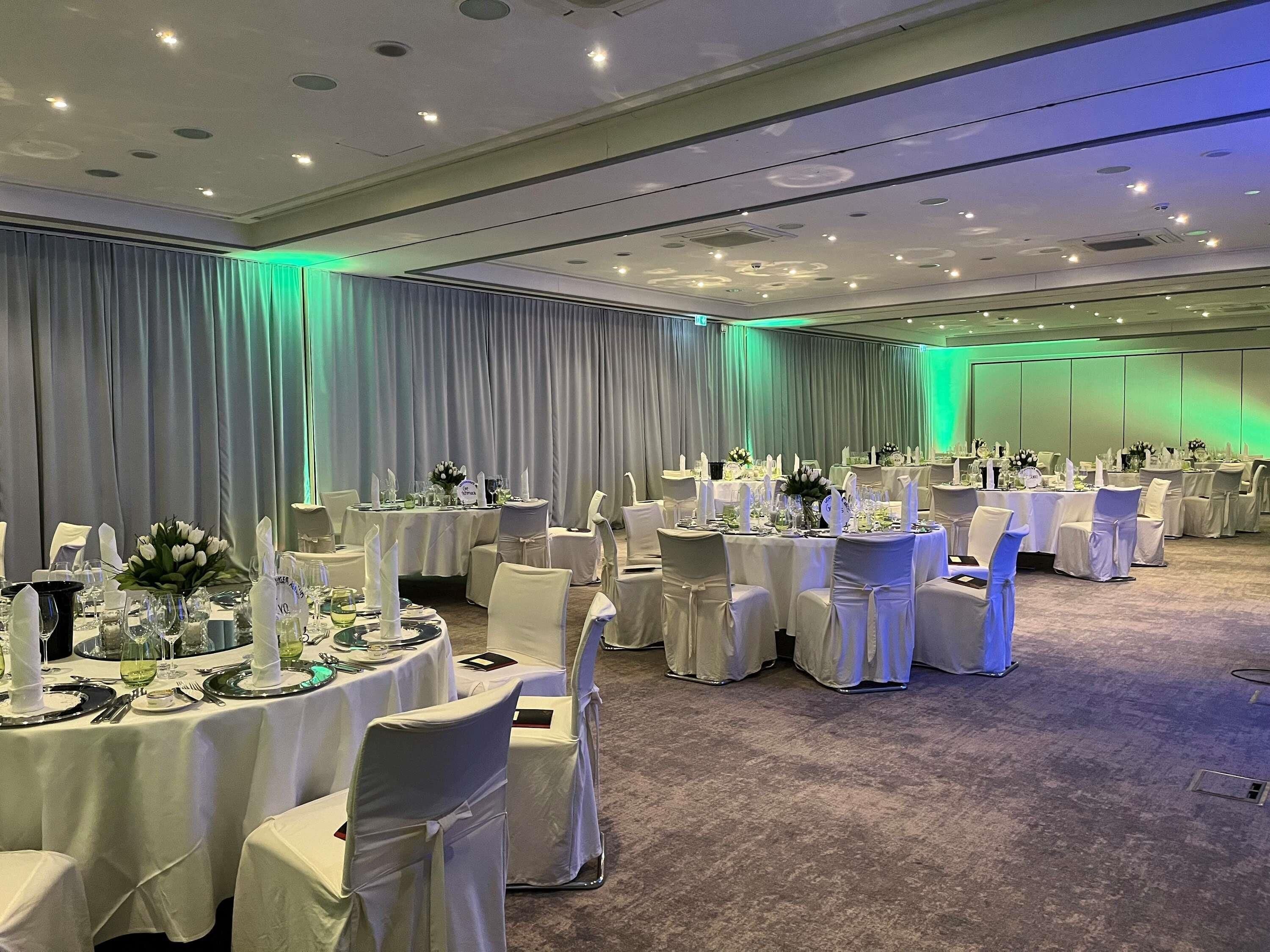 Un salón de banquetes con mesas redondas, manteles blancos y sillas con fundas blancas.