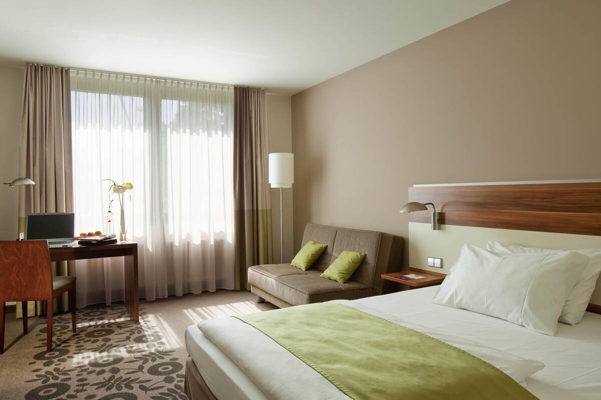 Habitación de hotel con una cama, un escritorio con una computadora portátil, una silla, un sofá y una mesita de noche con una lámpara.