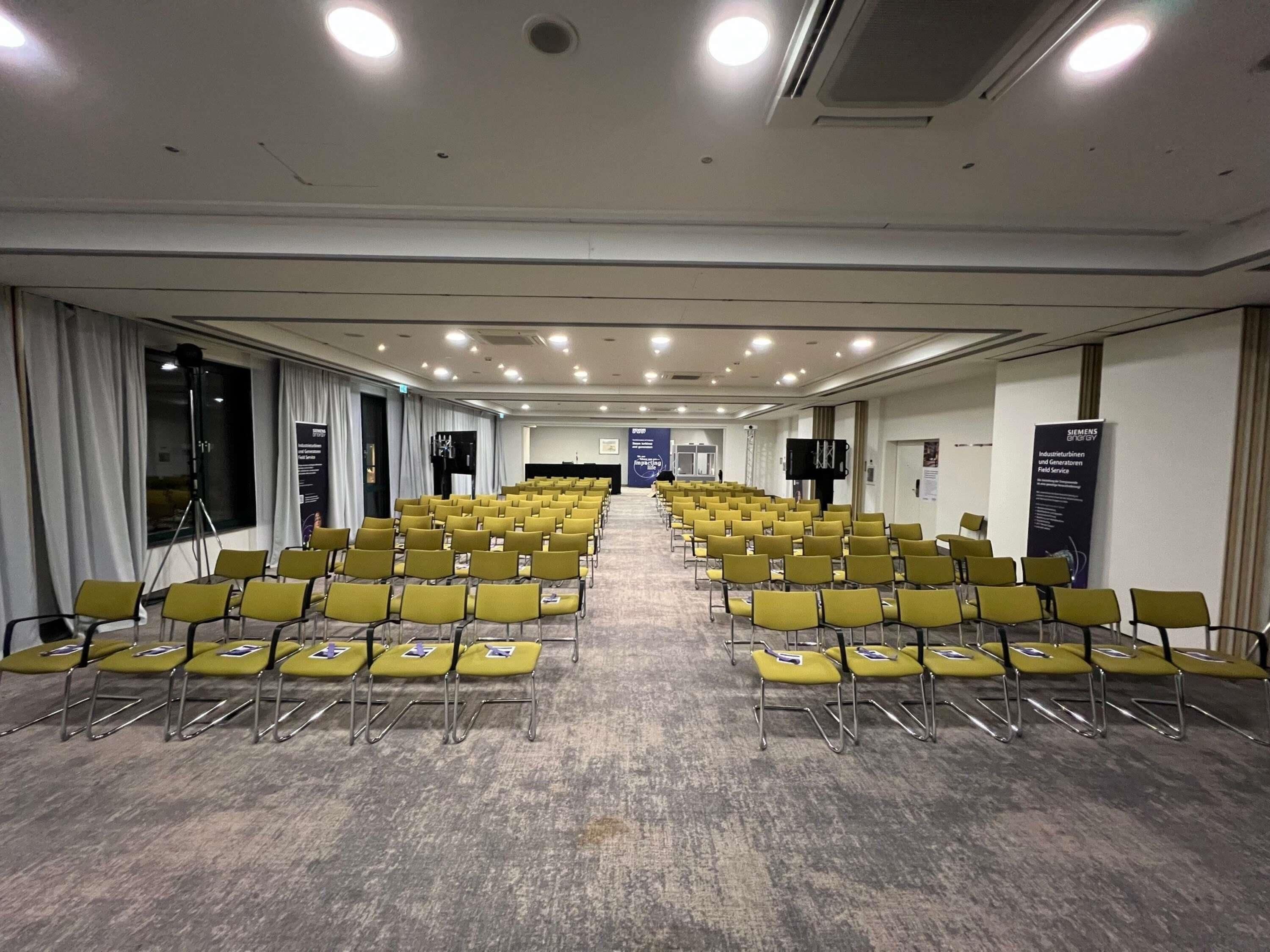 A conference room with rows of yellow chairs, a stage, and a banner on the wall.