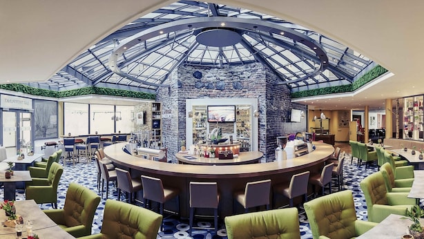 A hotel lobby with a central bar, green upholstered chairs, a patterned floor, and a glass ceiling. - Mercure Tagungs- & Landhotel Krefeld (Krefeld)
