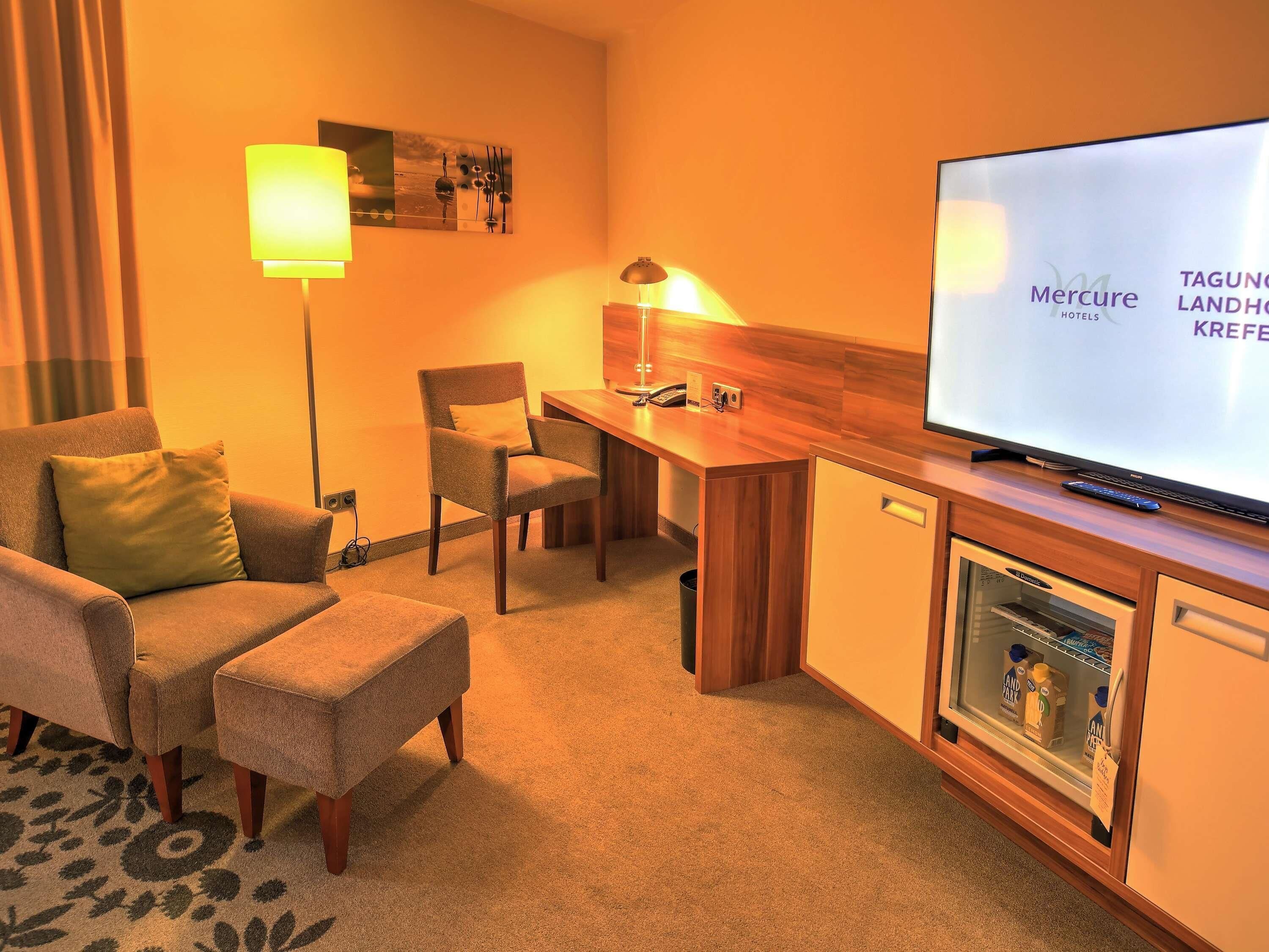 A hotel room with a sofa, armchair, a small table, a desk, a television, and a minibar.