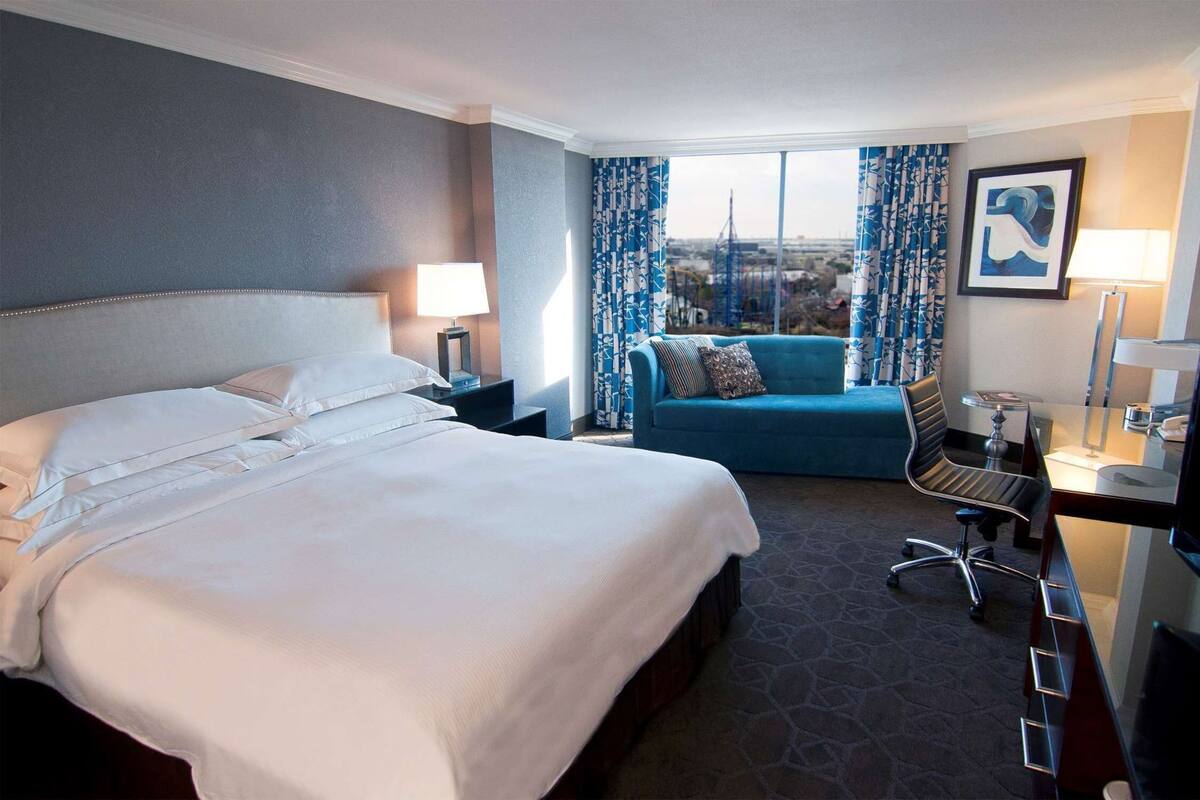 A hotel room with a large bed, a sofa, a desk with a chair, a TV, and a window with curtains.
