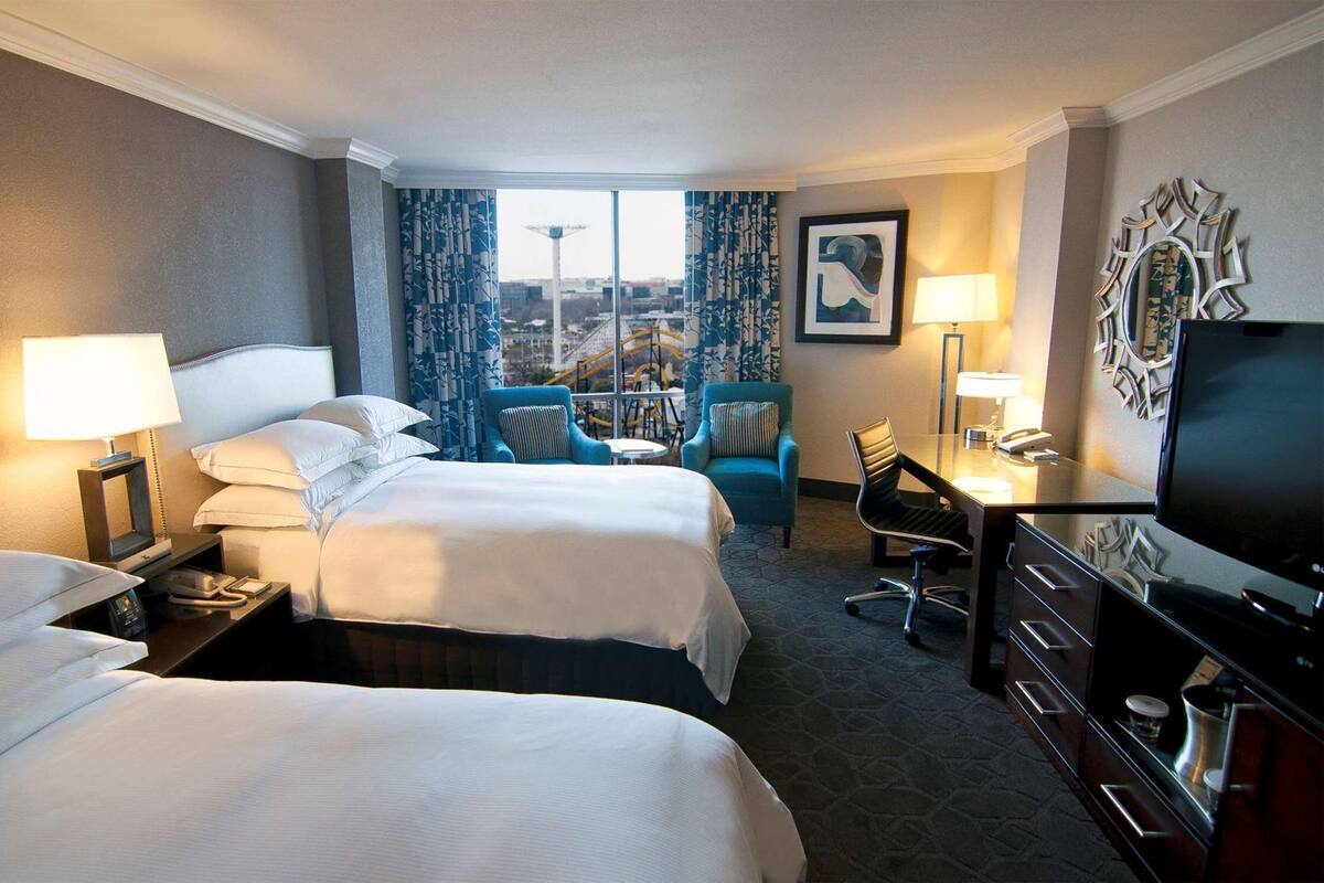 A hotel room with two beds, a desk with a chair, a TV, and a window with a city view.
