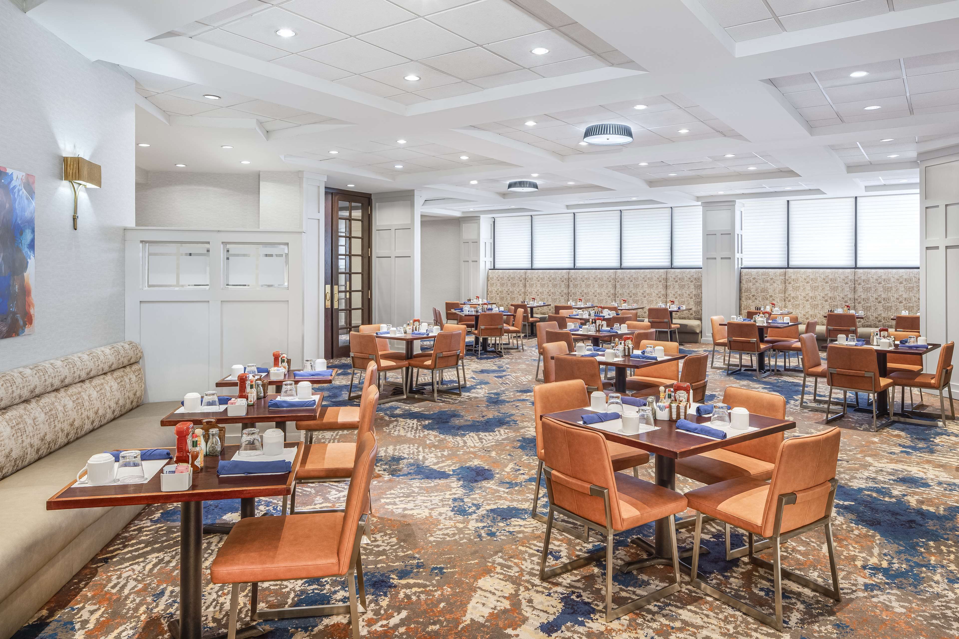 A hotel dining area with multiple tables and chairs, a patterned carpet, and a large window with blinds.