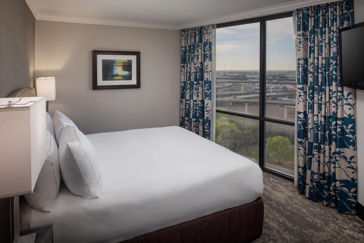 A hotel room with a large bed, a nightstand, a lamp, a framed picture on the wall, and a view of the city through a window with blue and white curtains.