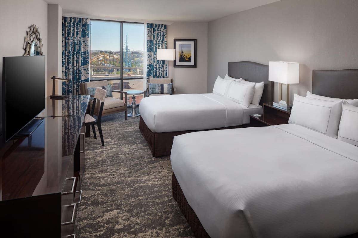 A hotel room with two beds, a desk, a chair, a TV, and a view of a cityscape.