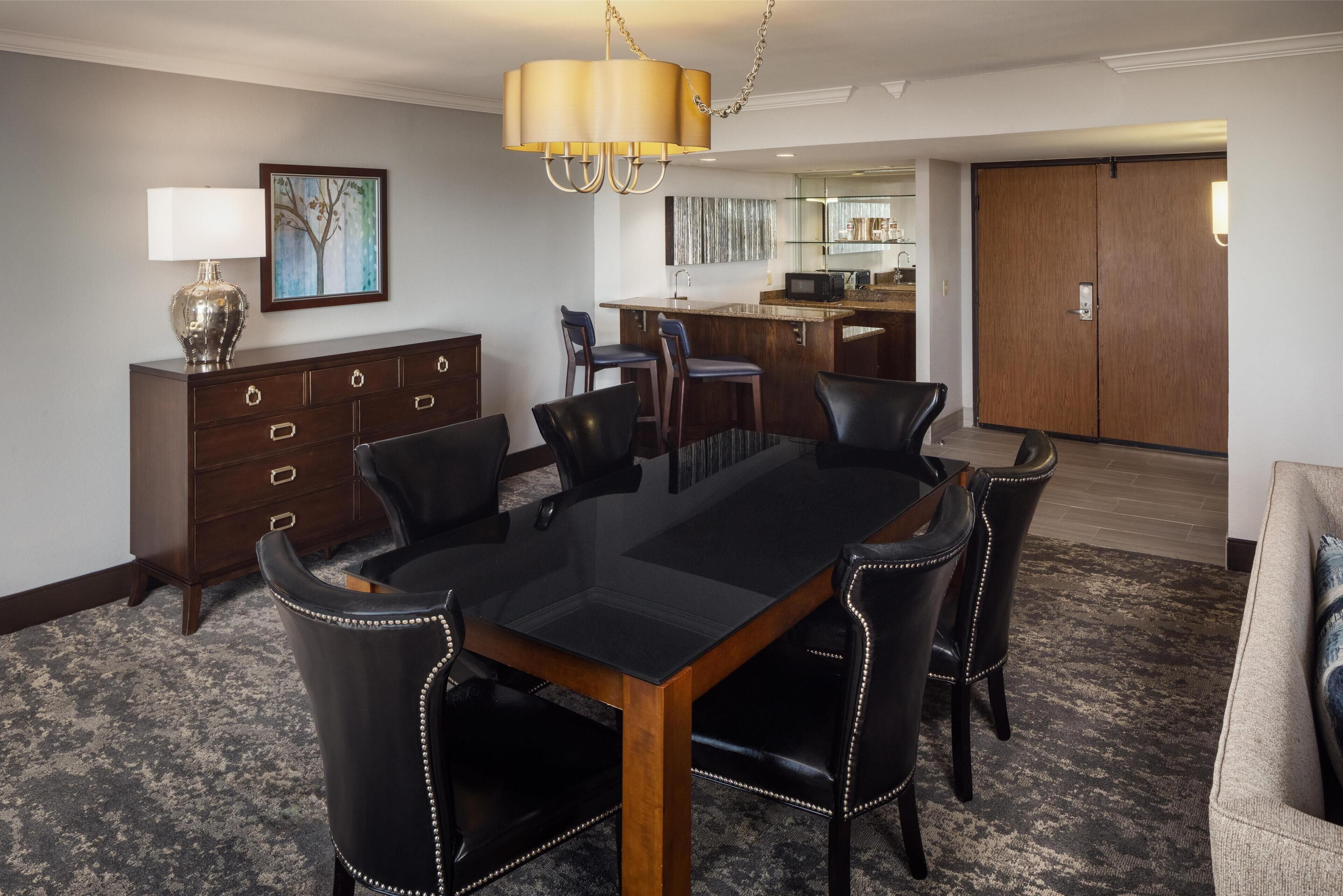 A hotel dining area with a large rectangular table, six chairs, a wooden cabinet, and a chandelier.