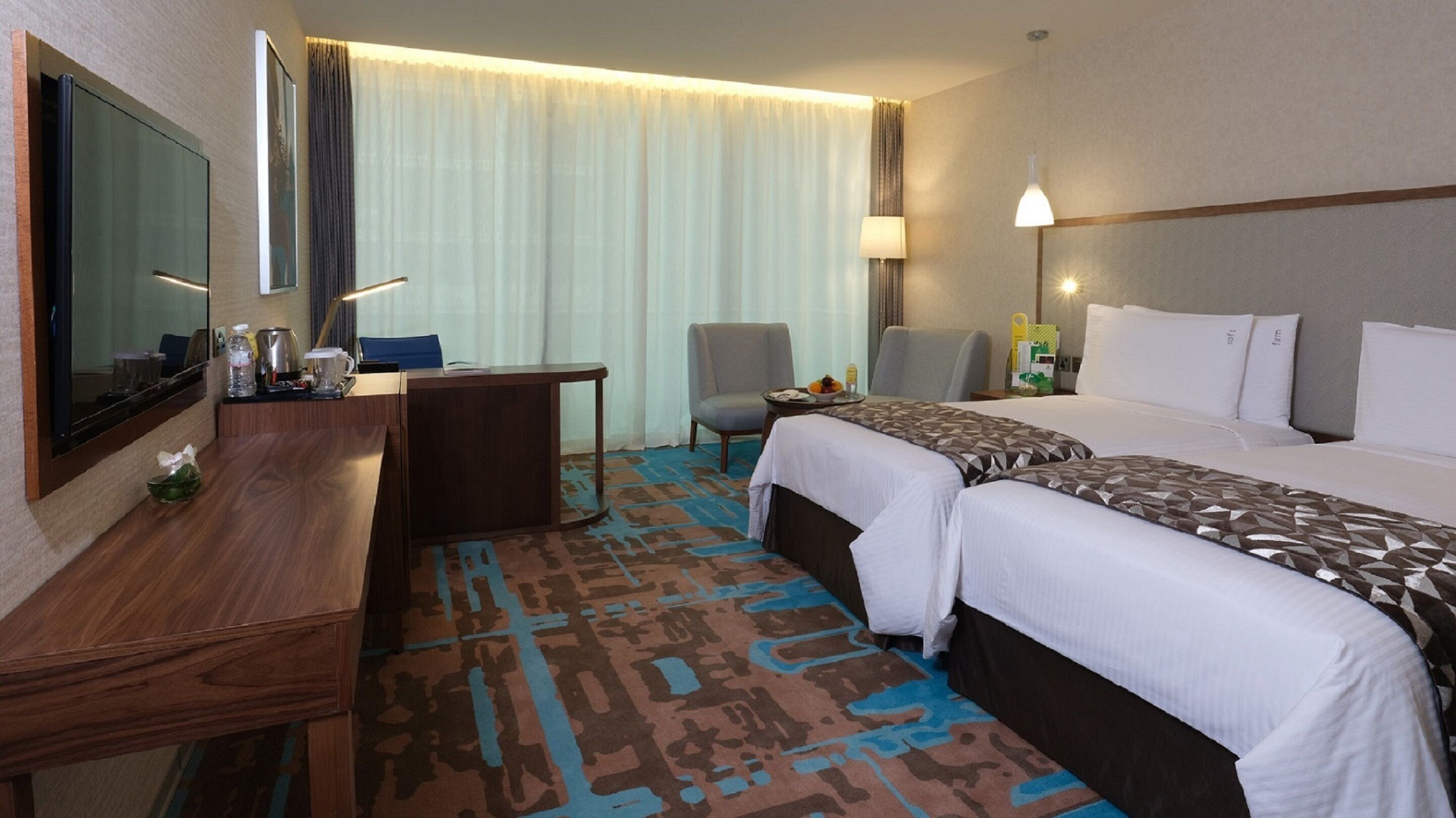 A hotel room with a desk, two beds, a chair, a lamp, and a large window with curtains.