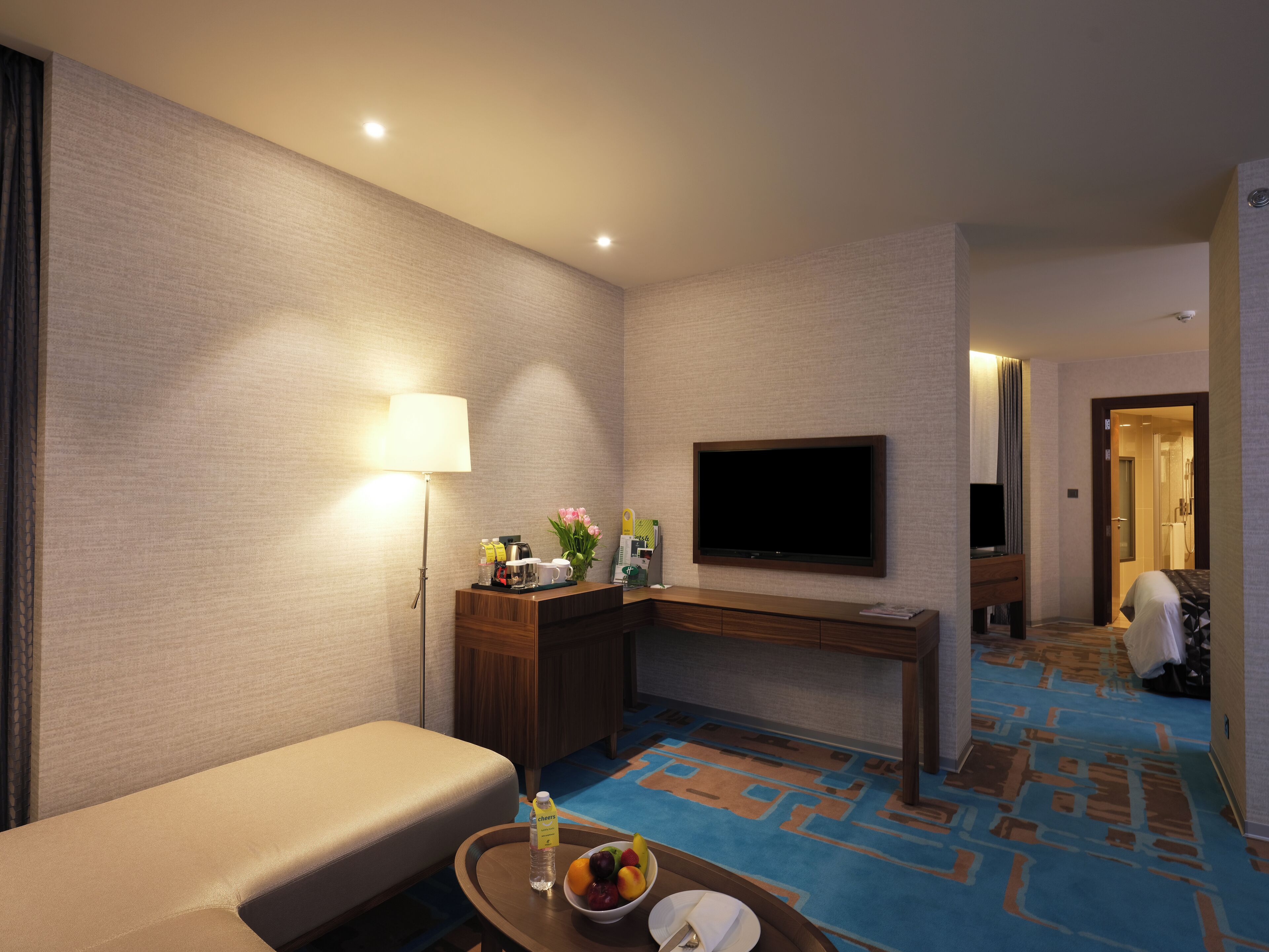 A hotel room with a sofa, a desk, a flat-screen TV, a lamp, a tray with a bottle and fruit, and a patterned carpet.