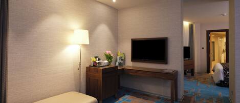A hotel room with a sofa, a desk, a flat-screen TV, a lamp, a tray with a bottle and fruit, and a patterned carpet.