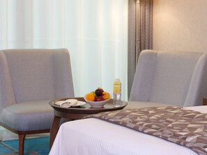A hotel room with two grey armchairs, a small round table with a fruit bowl, a bottle of water, and a bed with a patterned bedspread. - Holiday Inn Kuwait Al Thuraya City by IHG (Farwaniya)