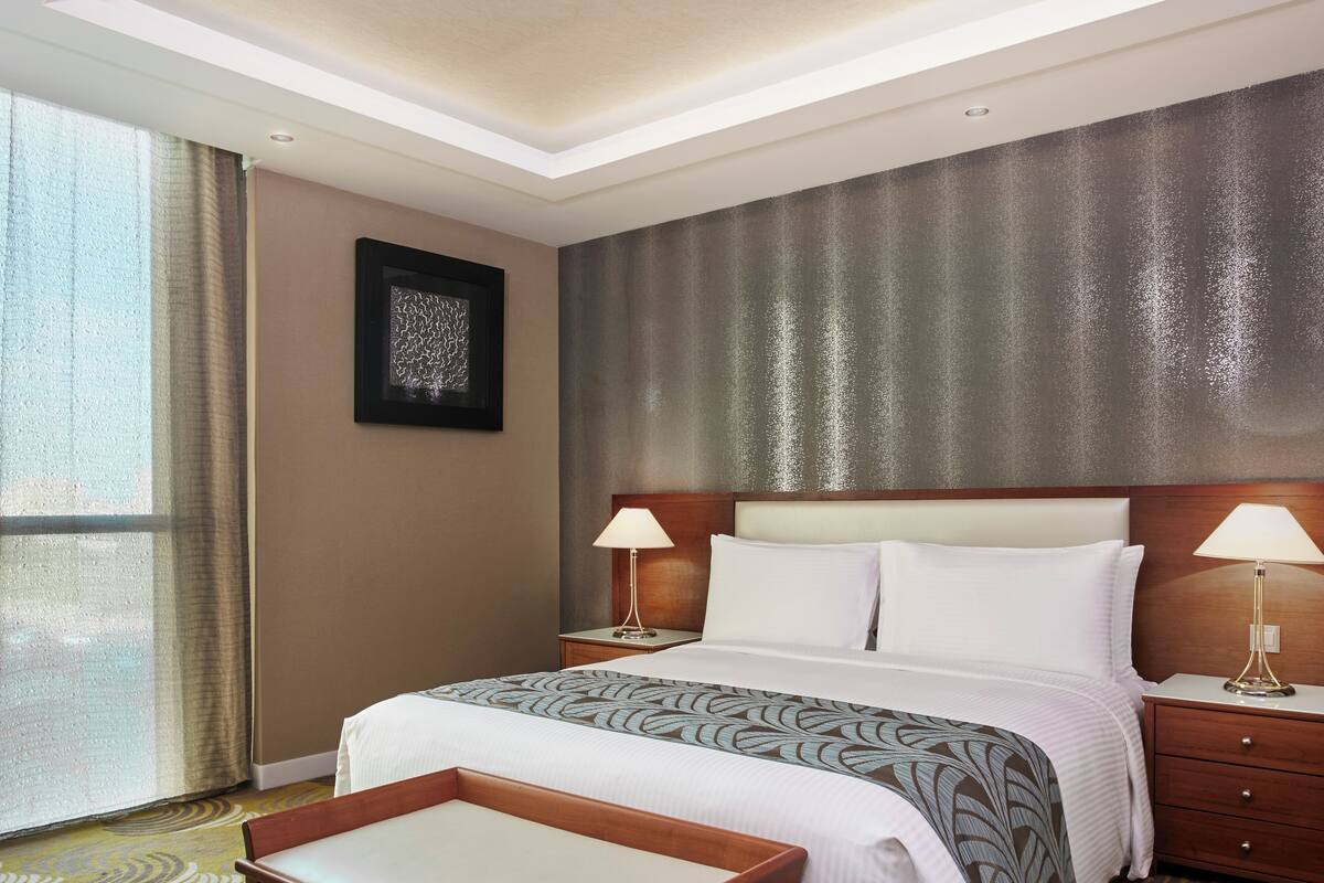 A hotel room with a large bed, two bedside tables with lamps, a small table, and a large window with curtains.