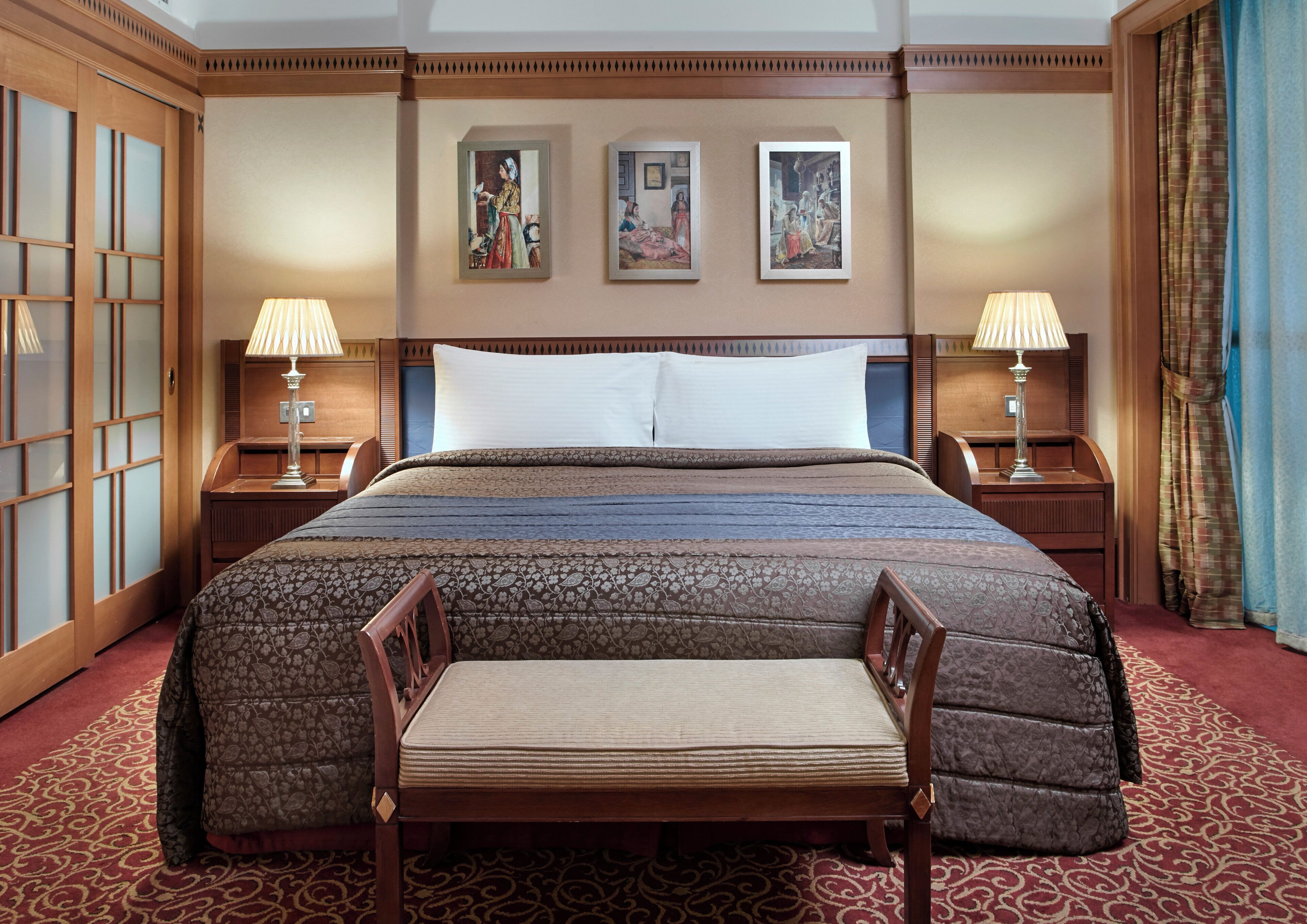 Habitación de hotel con una cama grande, dos mesitas de noche con lámparas, una silla de madera y una alfombra estampada.