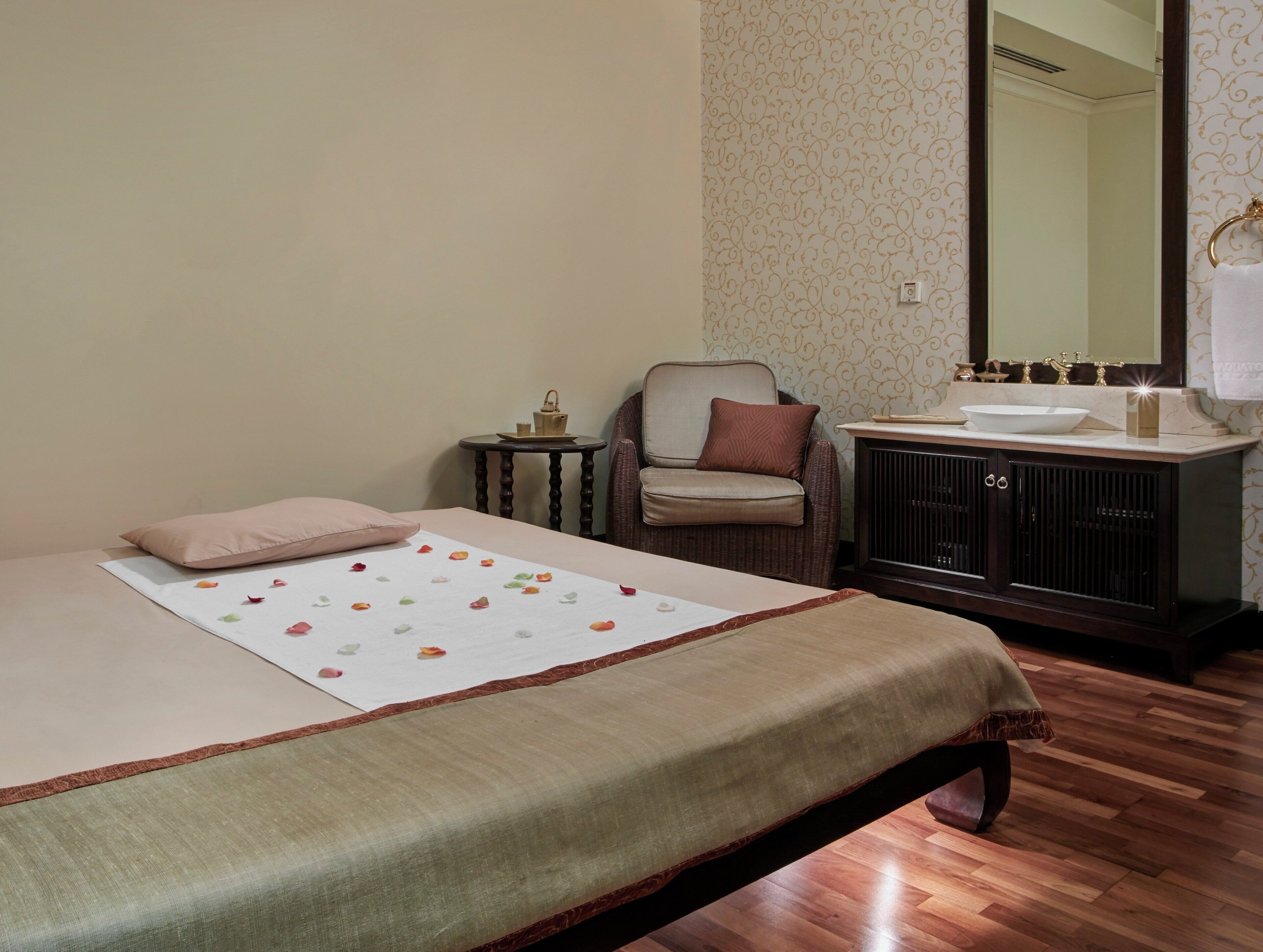 A bedroom with a bed, a chair, a small table with a lamp, a vanity with a mirror, and a wooden floor.